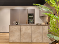 Moderne keuken met kookeiland in beige betonlook en matte kastfronten, voorzien van ingebouwde oven en koperen kraan.